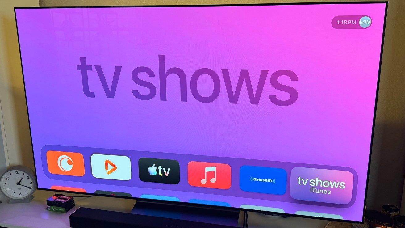 Large flat screen TV showing Apple TV home screen with pink purple background, tv shows heading, app icons row, and a small analog clock on the stand below.