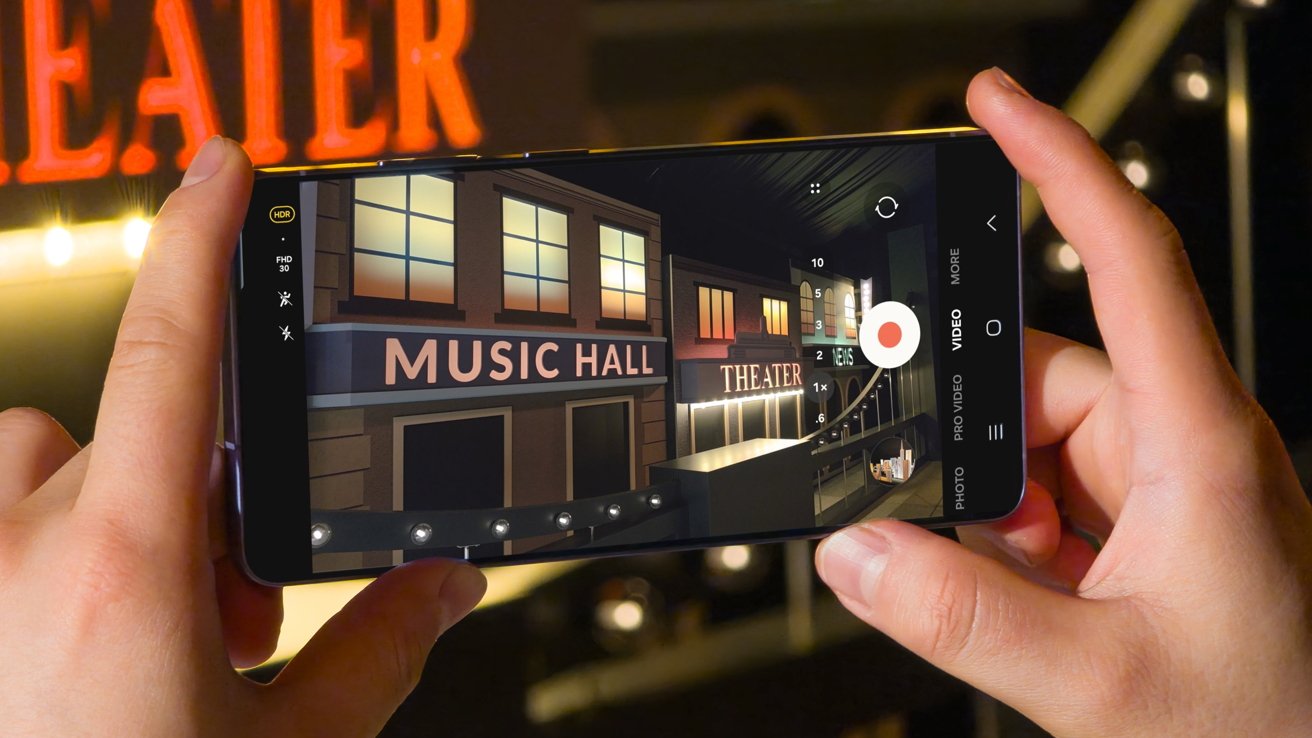 Hands holding a smartphone recording a nighttime street scene with bright signs reading MUSIC HALL and THEATER, warm glowing windows, and decorative lights in a theater district