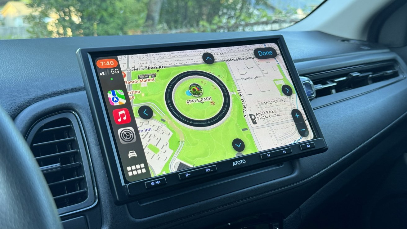 Car dashboard with large touchscreen displaying Apple CarPlay navigation map of Apple Park campus, including circular building, green surroundings, and app icons along the left side