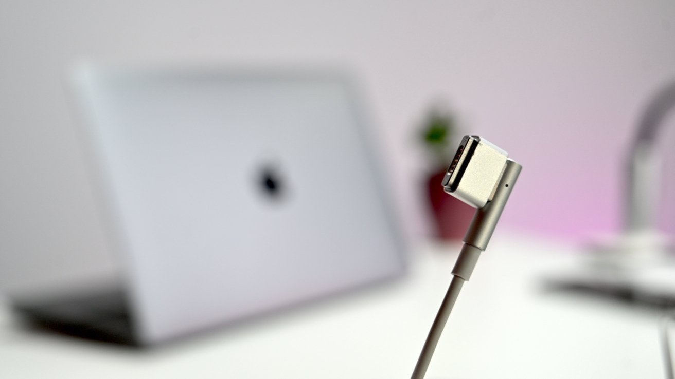 Close-up of a silver MagSafe-style laptop charging connector with a blurred open MacBook and soft pink-lit workspace in the background