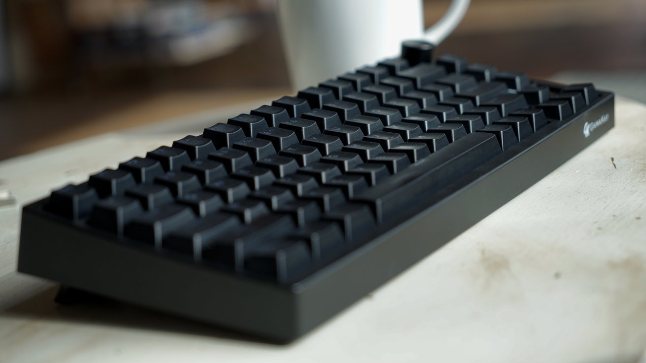 Compact black mechanical keyboard with tall keycaps on a light wooden surface, slightly angled, with a white coffee mug blurred in the background