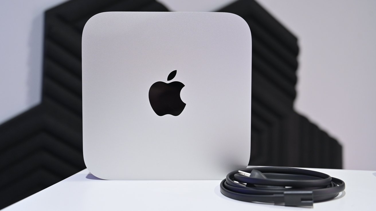 A silver Apple Mac mini desktop computer on a white surface with a black Apple logo, with a coiled black power or data cable lying to the right in front of a dark acoustic wall.