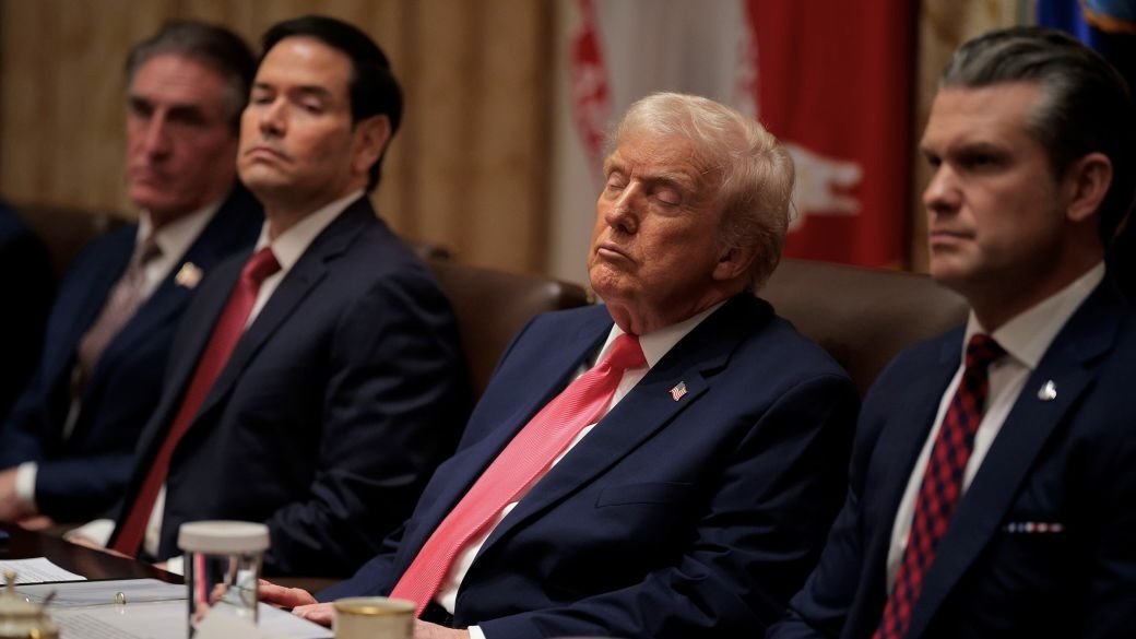 Four suited men sit at a conference table; the central man with blond hair and red tie has eyes closed, appearing contemplative, while others look forward in a formal meeting setting