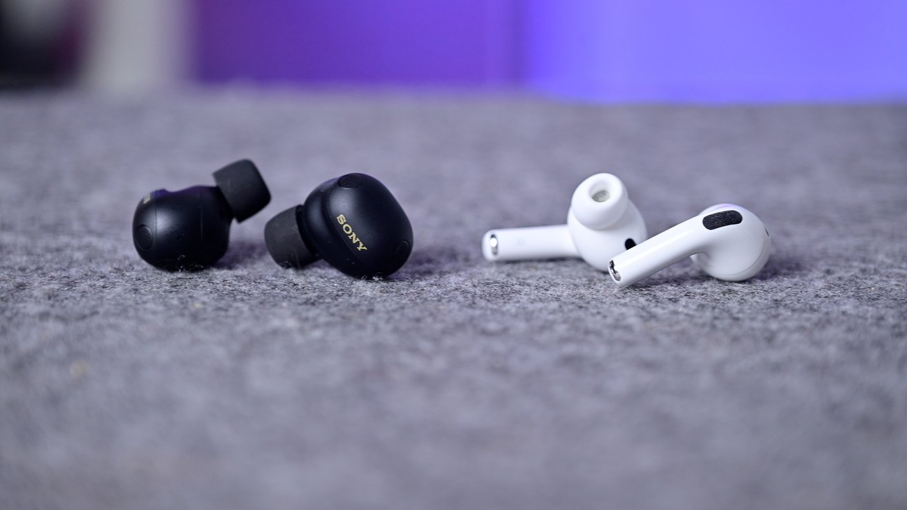 Two pairs of wireless earbuds rest on a gray fabric surface: compact black in-ear buds on the left and stem-style white earbuds on the right, against a soft purple background