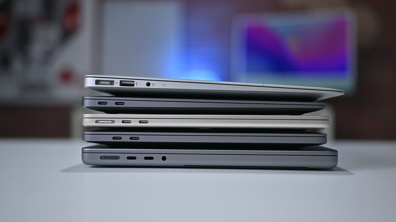 Stack of four closed laptops in different thicknesses and colors on a white table, showing side ports and slim designs, with a blurred, colorful office background