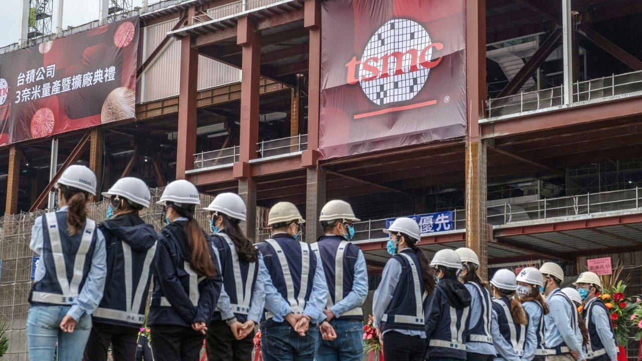 Construction workers wearing helmets and safety vests stand in a line facing a partially built factory with large TSMC banners and scaffolding in the background