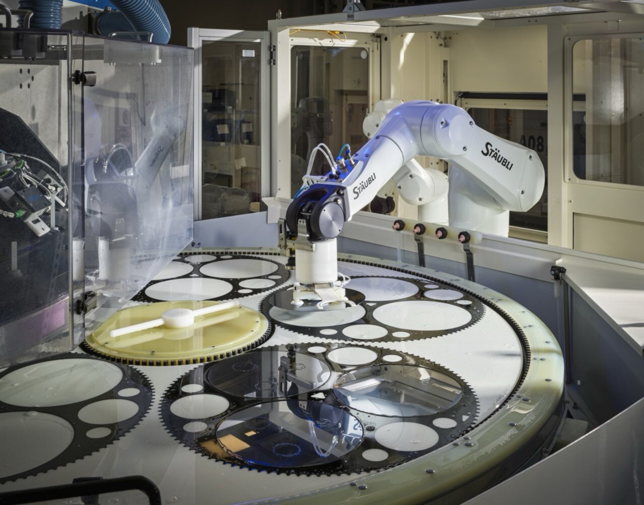 An industrial robotic arm working above a circular platform full of large interlocking gears and disks inside a clean, closed manufacturing environment with machinery and panels in the background