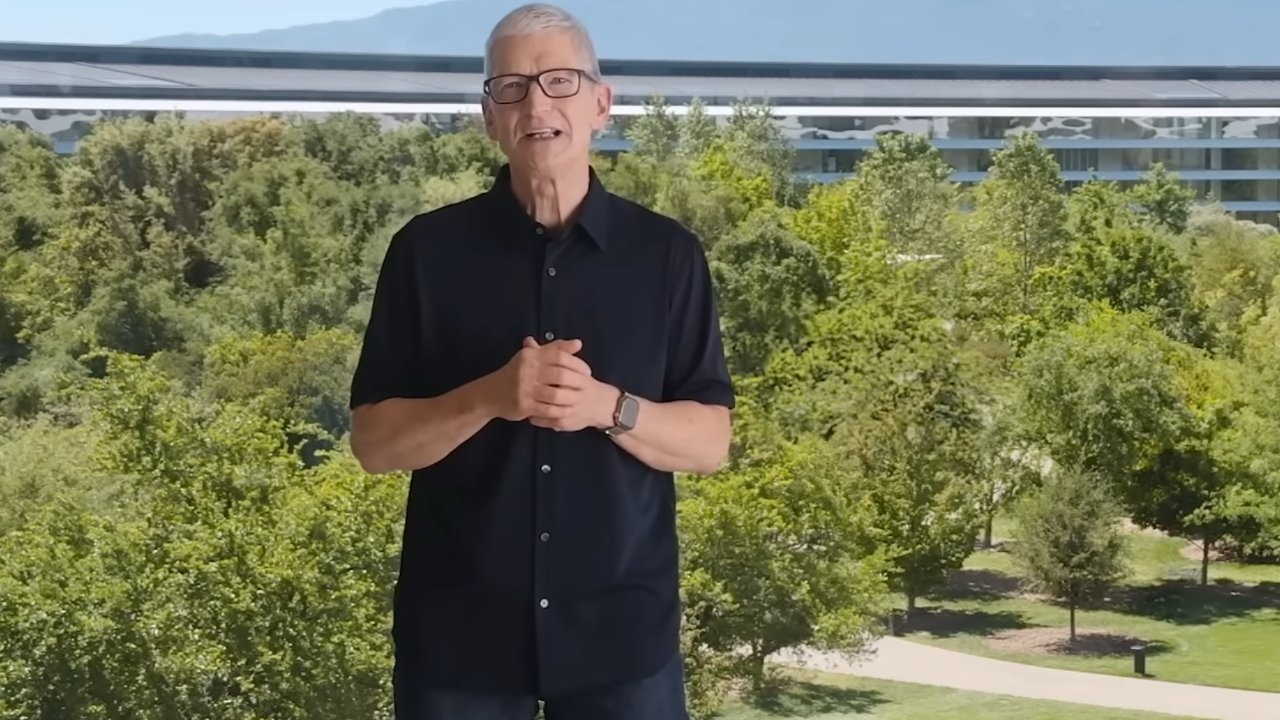 Older man in black shirt and glasses speaking outdoors, hands clasped, standing before lush green trees and a modern curved glass building in the background