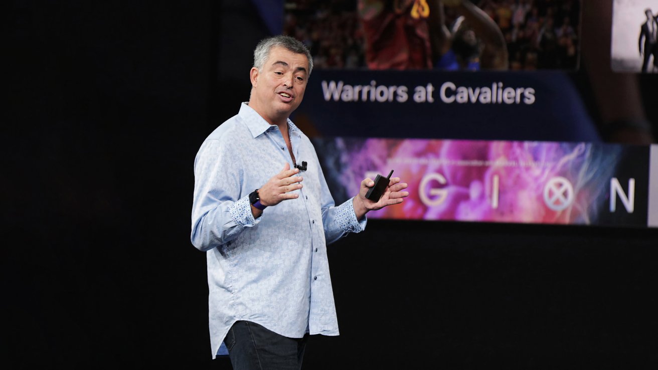 Middleaged man in a light blue shirt speaks onstage with a clicker, in front of a large screen showing Warriors at Cavaliers and colorful Legion graphics