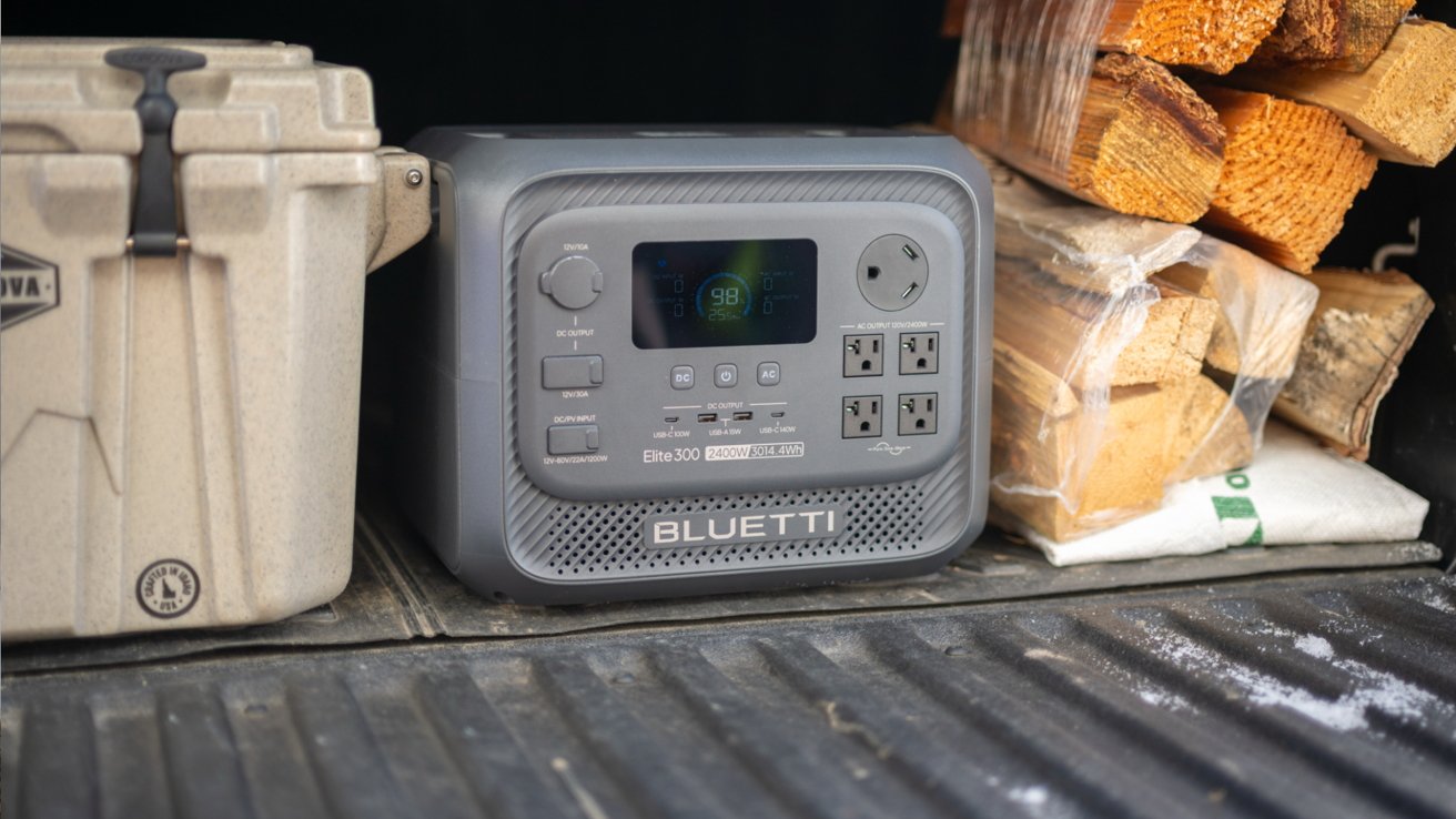 Portable Bluetti power station sitting in a truck bed between a rugged cooler and bundled firewood, showing multiple outlets and a lit digital display ready for outdoor use