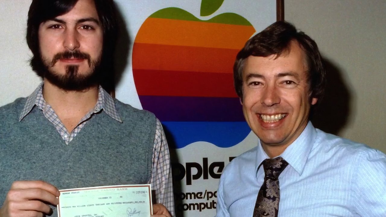 Two men standing and smiling in front of a large, colorful Apple logo; the bearded man on the left holds a check while the man on the right wears a tie