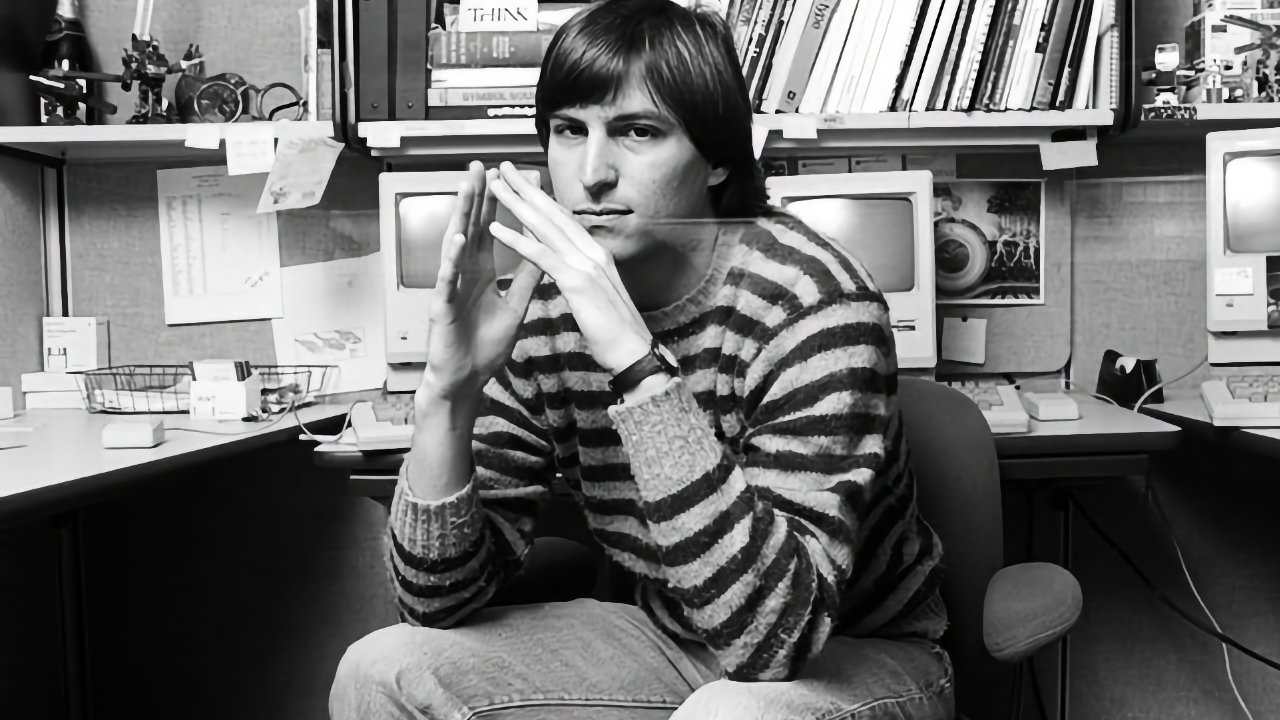 Young man in striped sweater sits thoughtfully in an office cubicle, hands steepled, surrounded by early desktop computers, papers, books, and shelves filled with binders and equipment