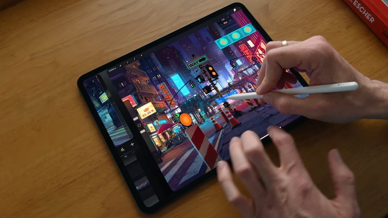 Hands using a stylus to digitally paint a colorful futuristic city street with neon signs and traffic barriers on a tablet resting on a wooden desk