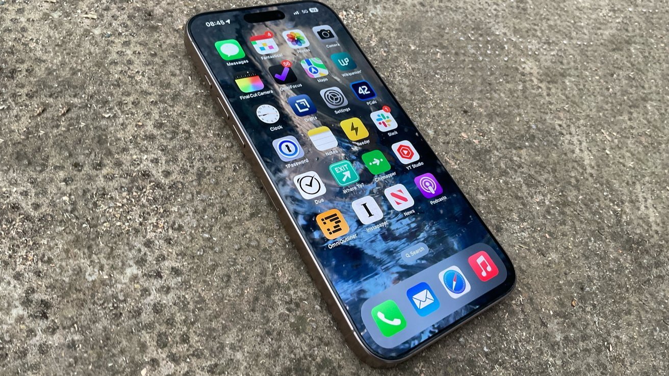Modern smartphone lying on rough concrete ground, screen on, showing a grid of colorful app icons and a reflective water-themed wallpaper.