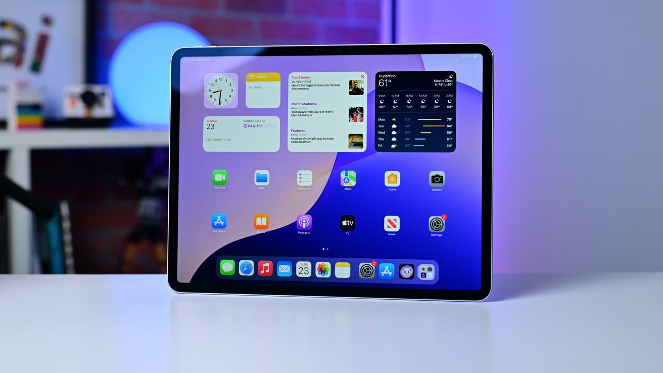 Tablet on a desk displaying colorful home screen with widgets, weather, and app icons; blurred background shows shelves and a glowing blue light sphere.