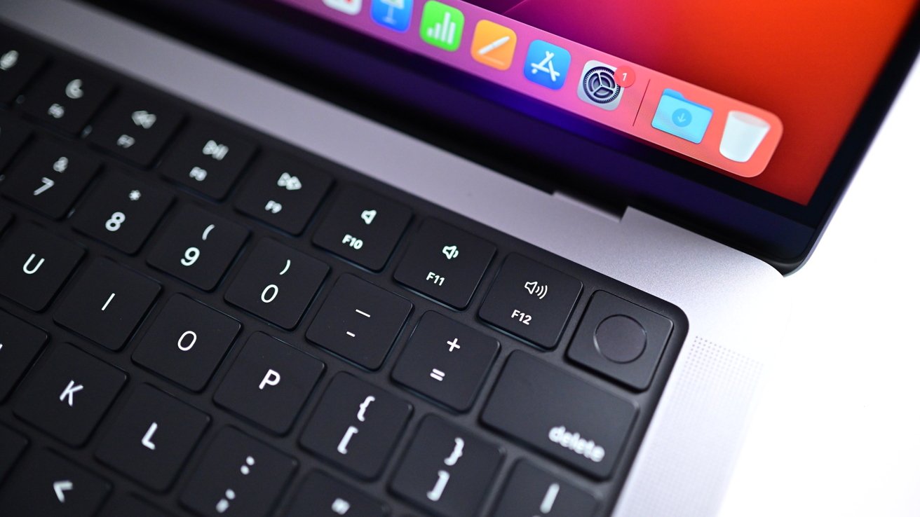 Close-up of a MacBook keyboard and trackpad area, showing black keys, volume function keys, Touch ID button, silver body, and part of the colorful macOS dock on the screen