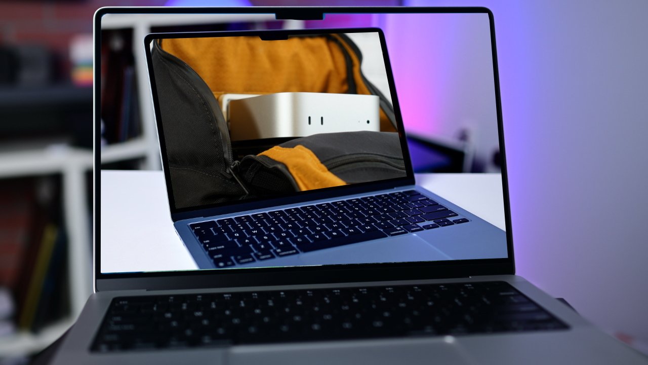 Open laptop on a desk, showing a photo of a backpack pocket holding a small silver device, with a blurred modern room and purple lighting in the background