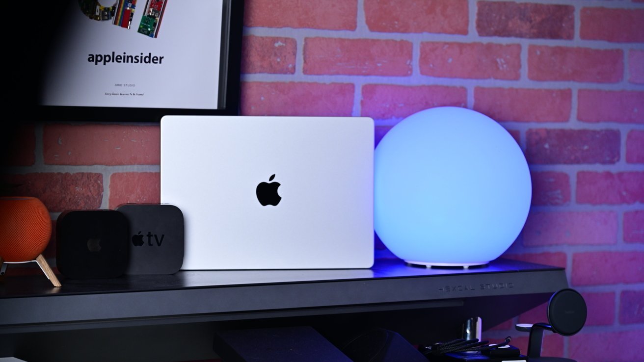 Silver MacBook with Apple logo on a desk, flanked by Apple TV boxes, orange smart speaker, glowing blue spherical lamp, and framed appleinsider poster against a red brick wall