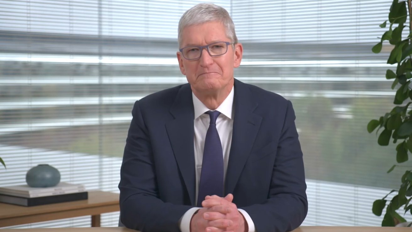 Older man with short gray hair, glasses, and navy suit sits at a desk, hands clasped, in a bright office with window blinds and potted plants in the background