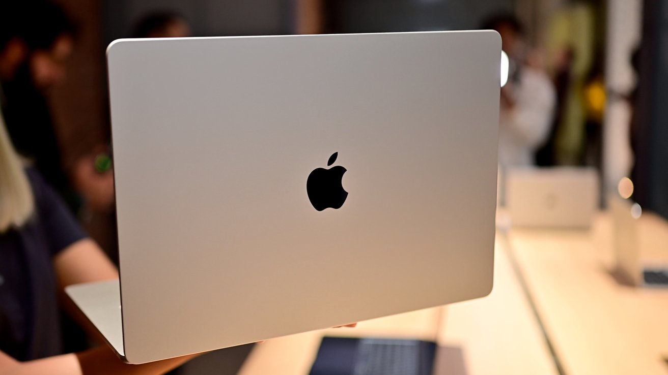 Person holding a silver Apple laptop by its edge, lid open away from camera, showing large black Apple logo centered on the smooth metallic back, with blurred people and tables behind