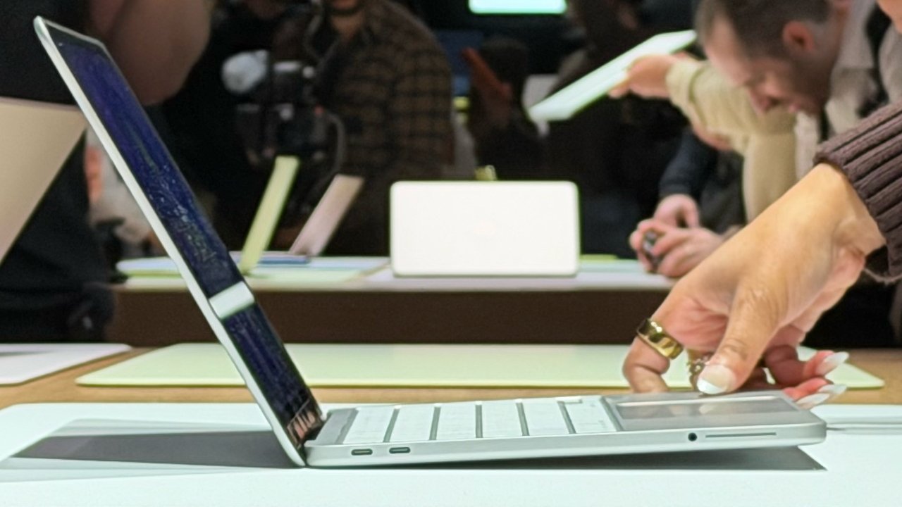 Person testing a slim laptop on a table, touching its trackpad, with other open laptops and people interacting with devices in a busy, well-lit tech demonstration setting