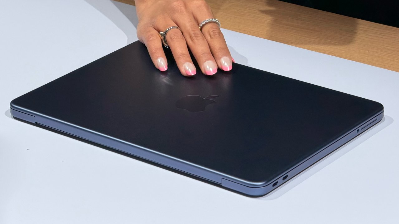 Hand with pink manicured nails and silver rings resting on a closed dark blue Apple MacBook on a white surface with a wooden table edge in the background