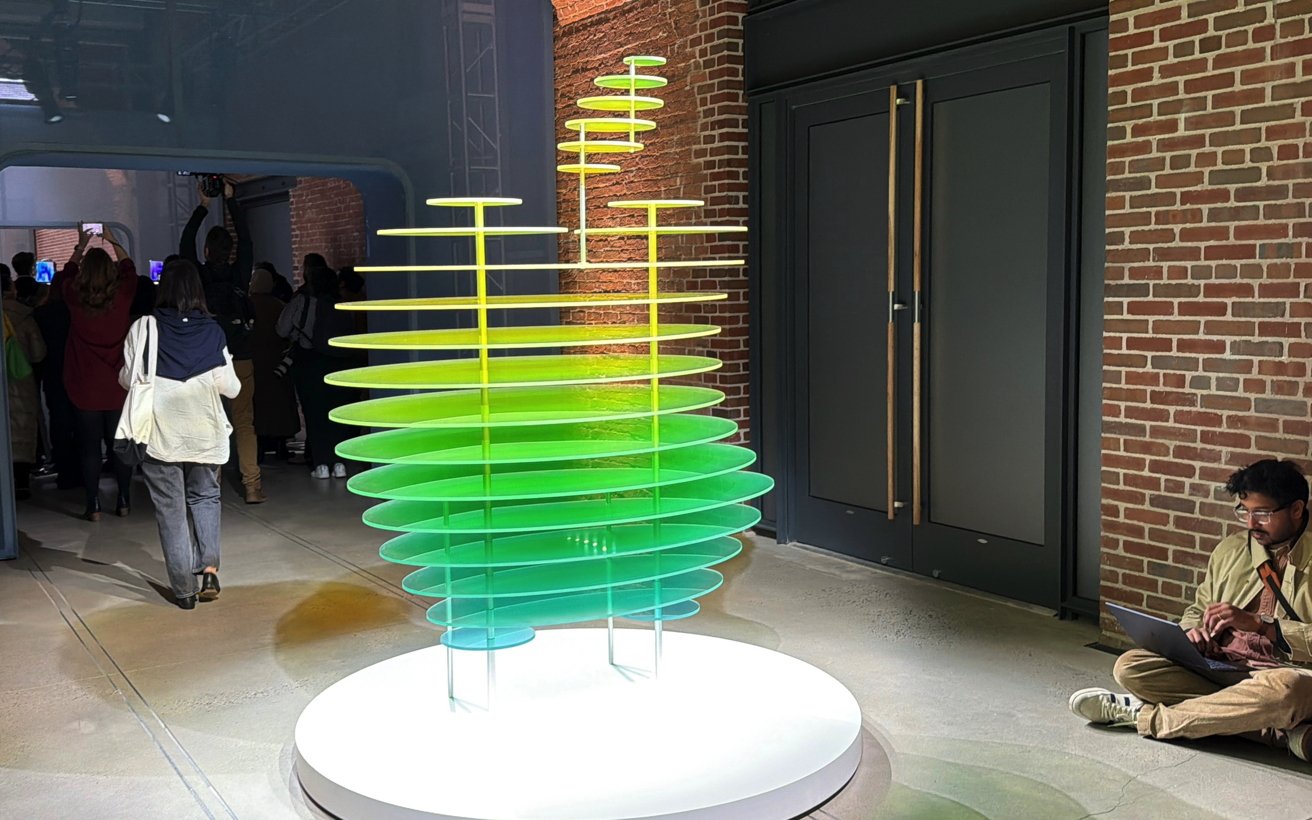 Colorful layered Apple logo glass sculpture glowing green and yellow on a round pedestal in an industrial gallery, with visitors in the background and a man sitting on the floor using a laptop