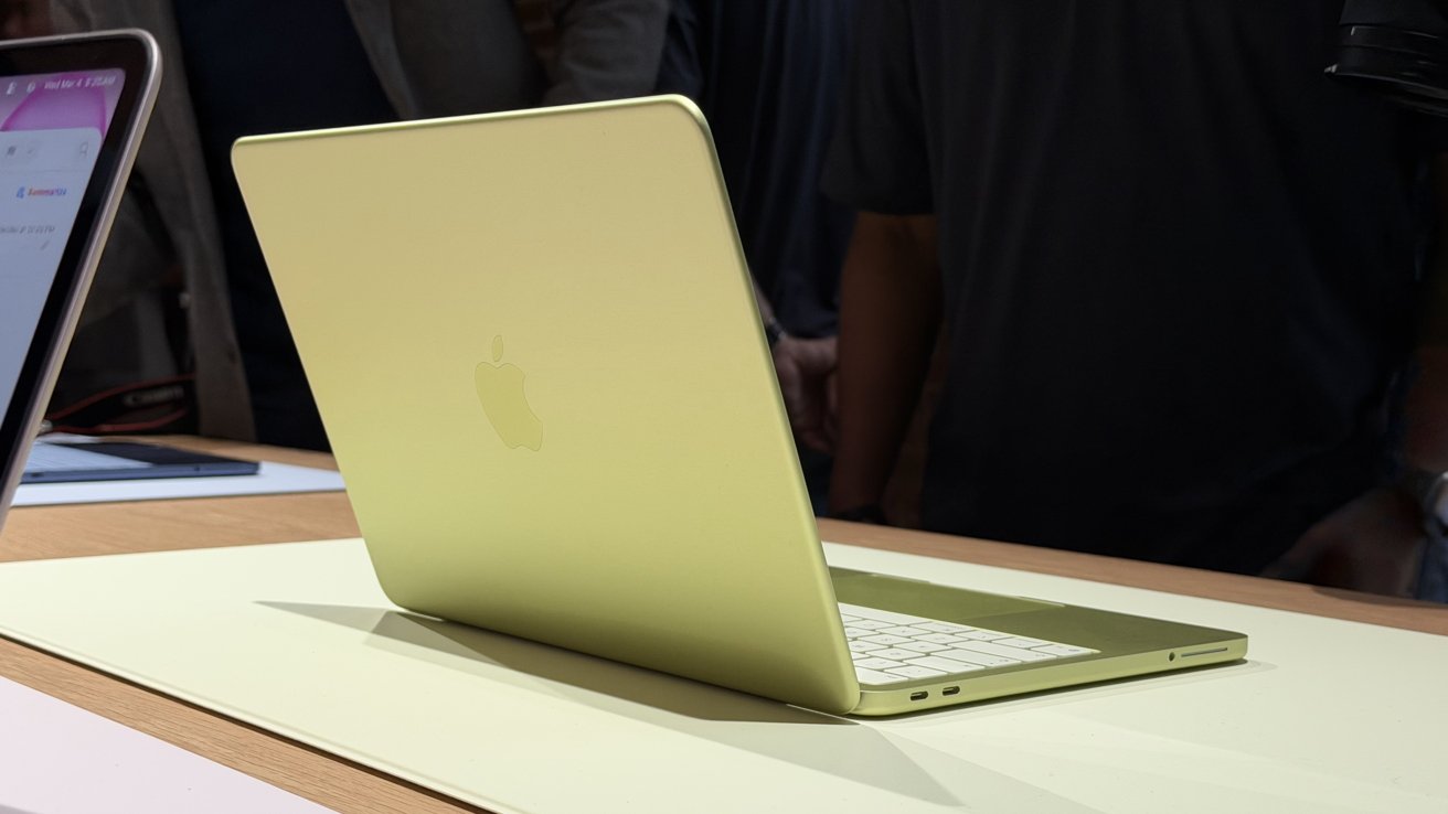 Open yellow Apple laptop on a light desk, viewed from behind, with white keyboard, slim ports on the side, and people standing in the dim background