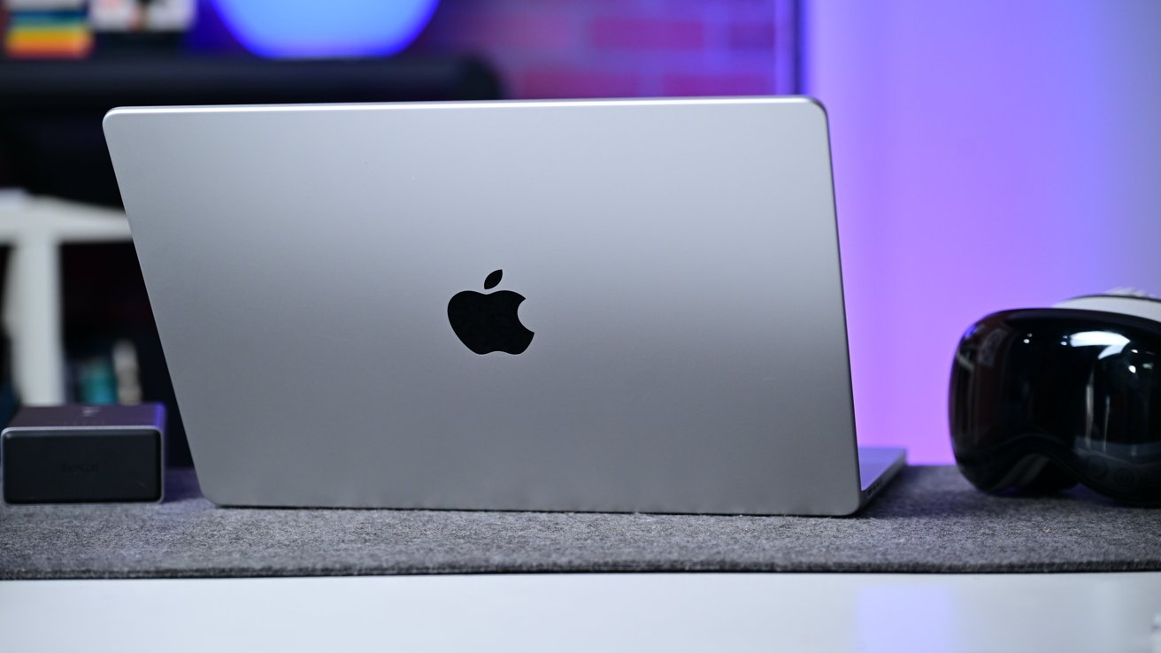 Silver MacBook with Apple logo closed on a desk mat, VR headset to the right, small black device to the left, blurred colorful background lighting