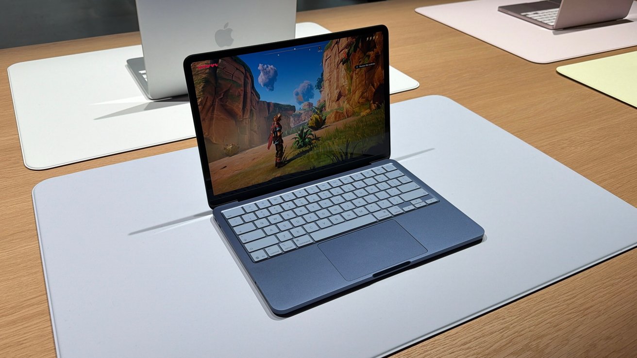 Blue Apple MacBook Neo on display table, screen showing colorful canyon video game scene with character; other open MacBooks in soft colors sit blurred in background on wooden surface
