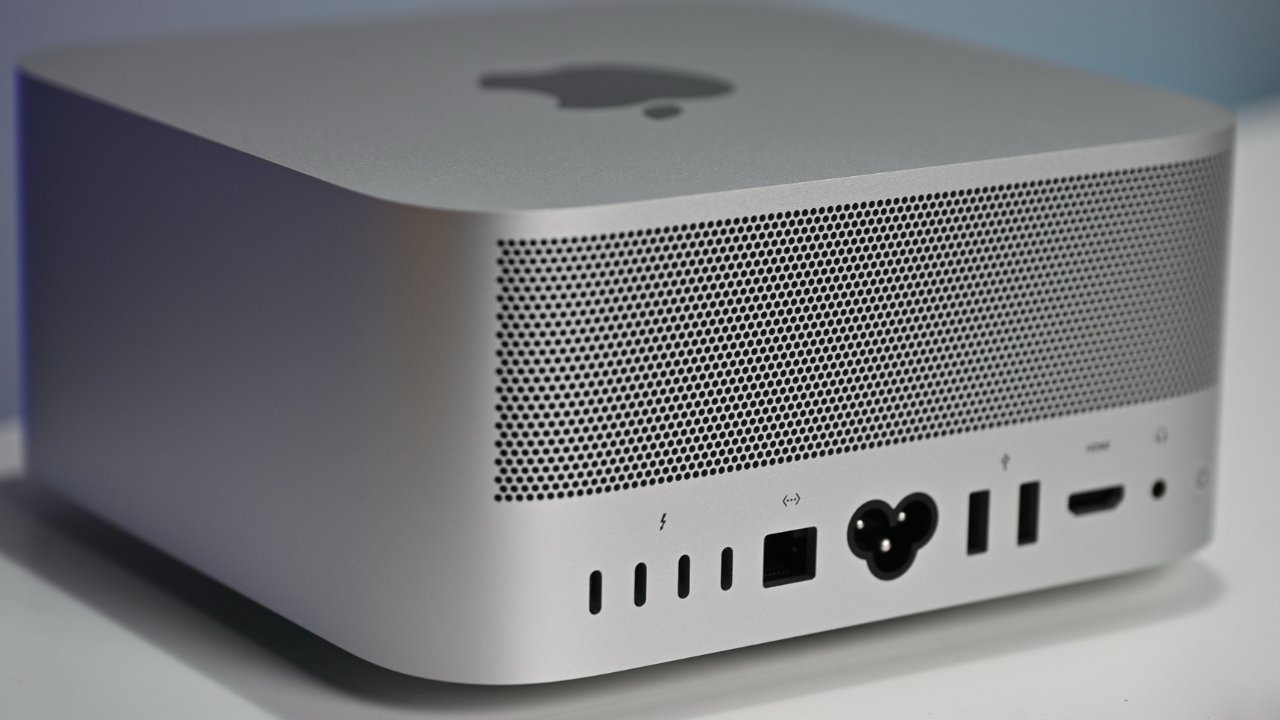 Silver Apple desktop computer with perforated rear panel, showing power connector, USB, Thunderbolt, Ethernet, HDMI, and other ports, placed on a light surface with soft lighting