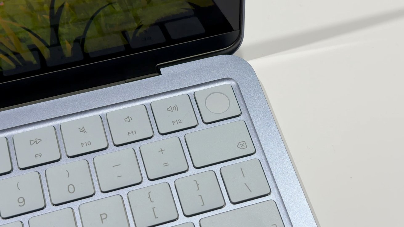 Close-up of a silver laptop keyboard showing function keys F9-F12, volume controls, a large circular power or fingerprint button, and part of the hinge and screen edge on a white surface