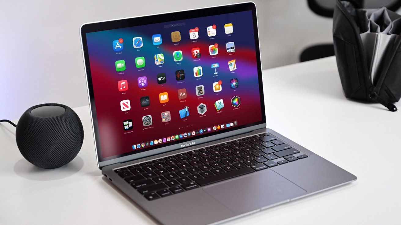 Open MacBook Air on a white desk showing colorful app icons, with a small black smart speaker on the left and a black organizer bag blurred in the background