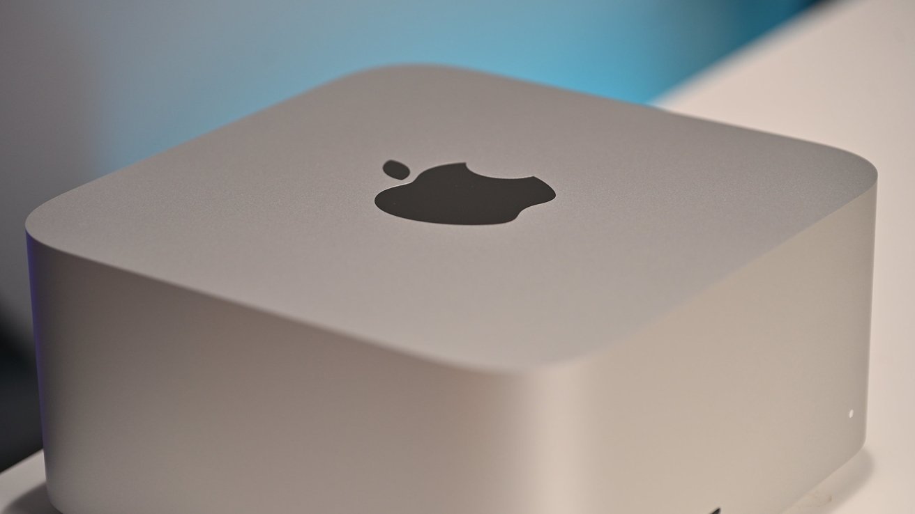 Silver Apple Mac mini desktop computer with rounded corners, viewed close up from above, showing the black Apple logo on top against a softly lit blurred background