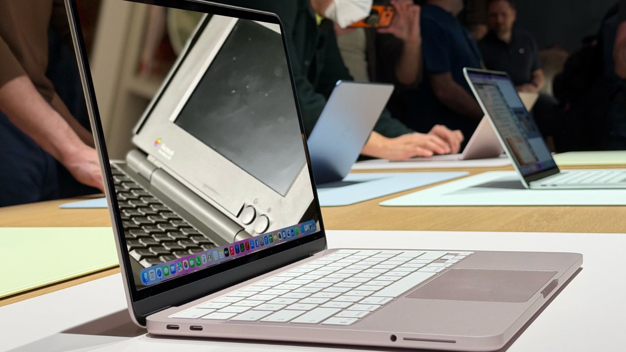 Several thin Apple laptops on a table at a tech event, with the closest screen showing a photo of an old Macintosh notebook while people in the background test other computers