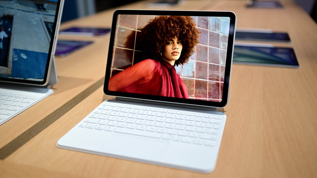 Tablet with white keyboard on a wooden table, displaying a woman with curly hair in a red outfit standing against a tiled wall in a modern electronics store