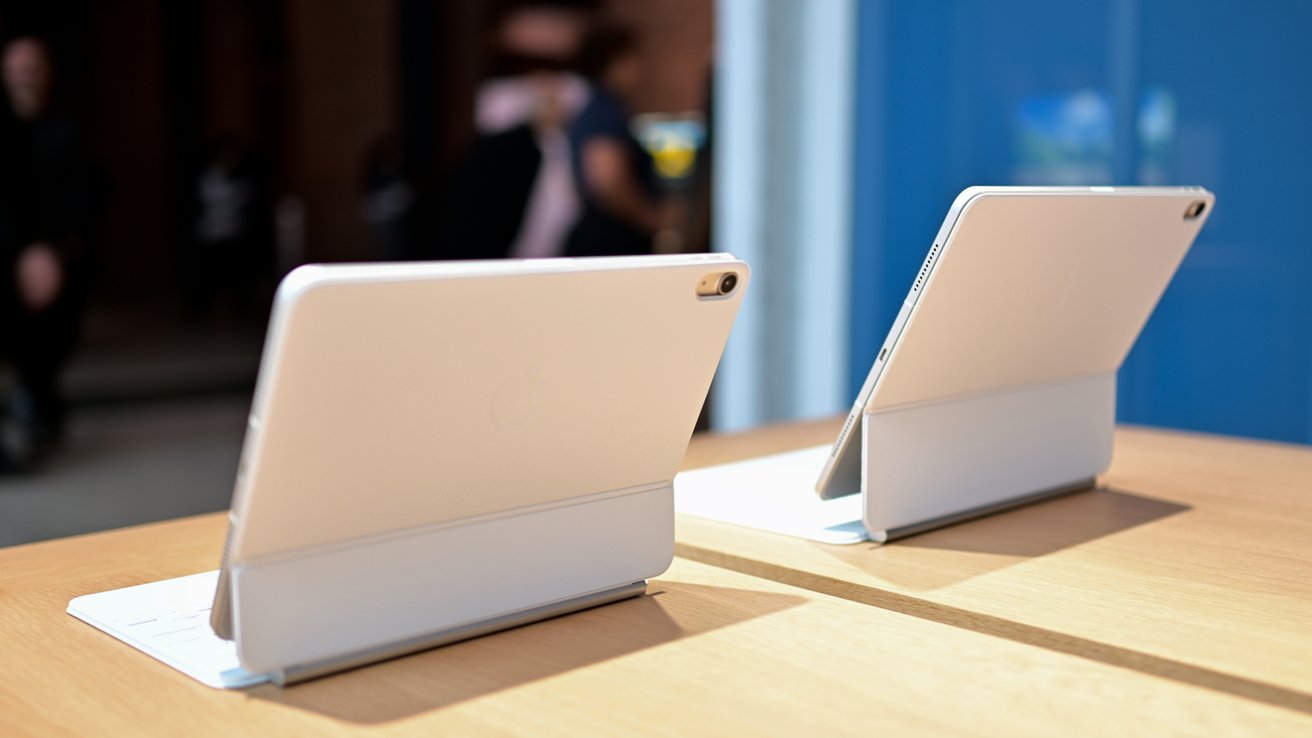 Two slim white tablets on stands sit on a wooden table in a bright store, facing away from the viewer, with blurred people and a blue wall in the background