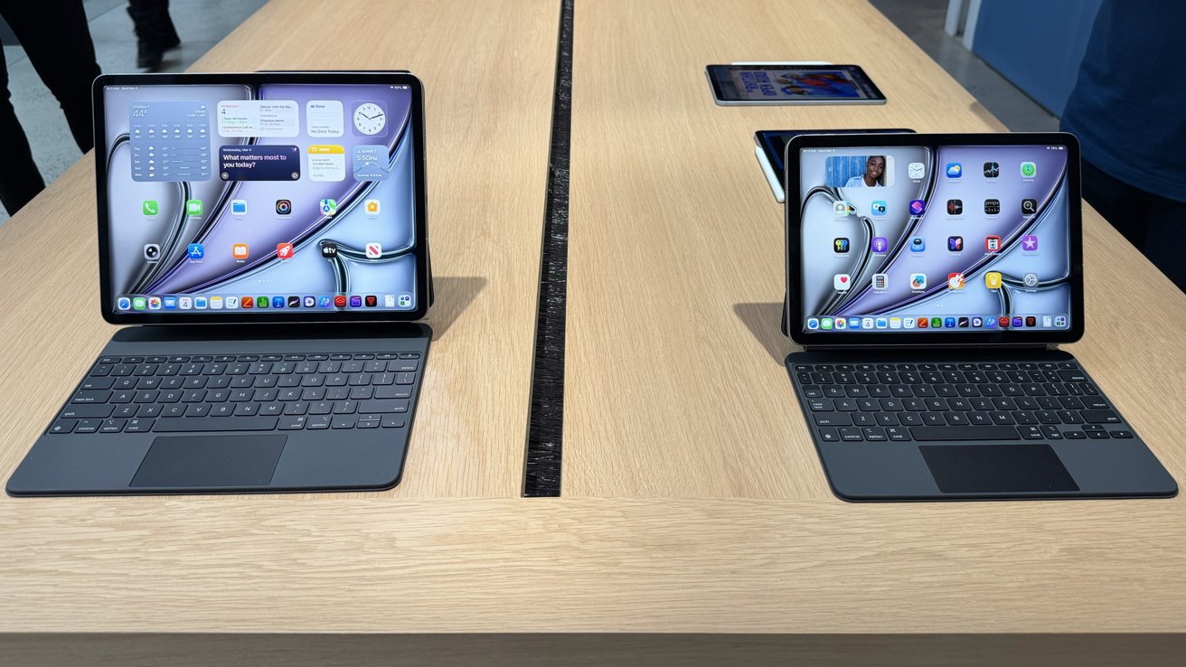 Two iPad tablets on a wooden store table, each attached to a black keyboard case and displaying colorful home screens; additional tablets are visible further back on the table