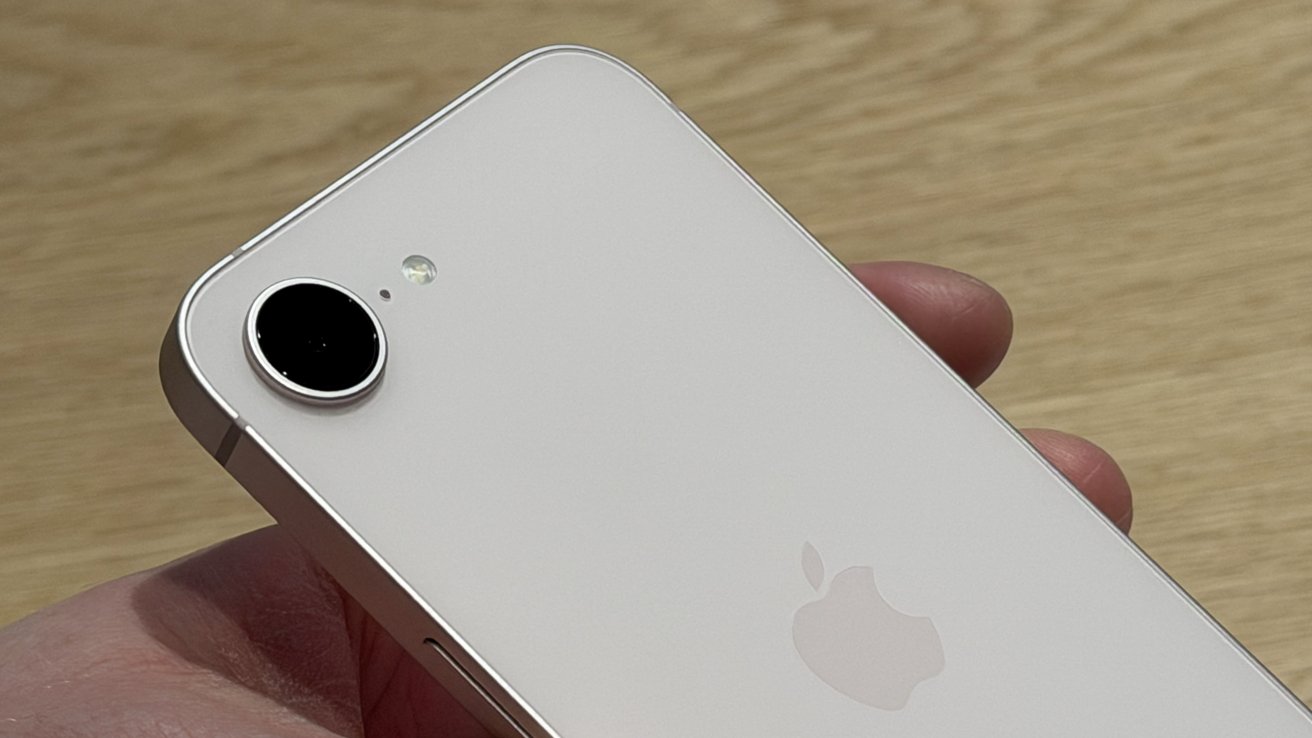 Hand holding a white iPhone, showing the back with a single rear camera, flash, and Apple logo against a light wooden background
