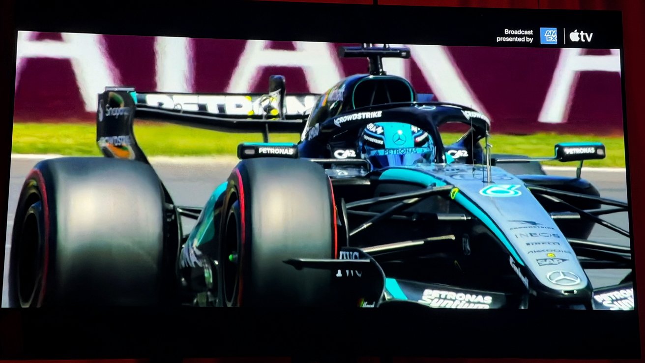 Close-up of a Mercedes Formula 1 car racing on track, driver in blue helmet, large slick tires, teal and black livery, sponsorship logos visible, blurred background barriers and grass.