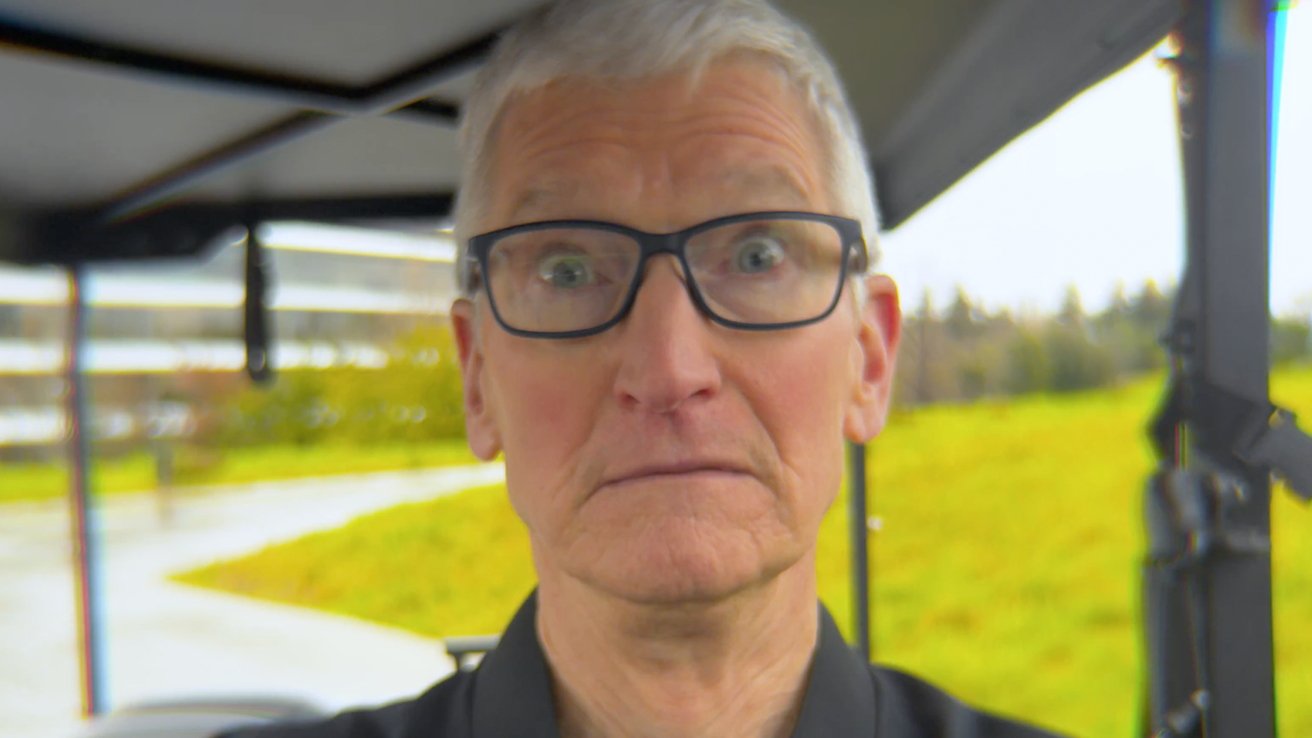 Older man with short gray hair and glasses staring wide-eyed at the camera, standing outdoors near green grass and modern buildings on an overcast day