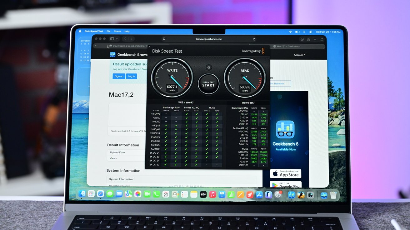 Open MacBook on a desk running a disk speed test and Geekbench benchmark in Safari, showing performance dials and result tables against a blurred purple and blue background