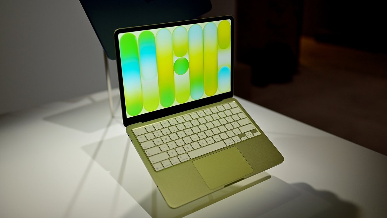 Slim light-green laptop on a stand, with white keyboard and large trackpad, displaying a colorful abstract wallpaper of vertical rounded shapes in green, yellow, and blue tones.
