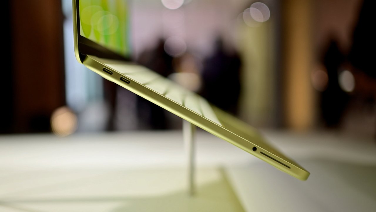 Side view of a thin, gold-colored laptop on display stand, lid open, showing slim keyboard, two USB-C ports, headphone jack, and blurred indoor background with soft lights