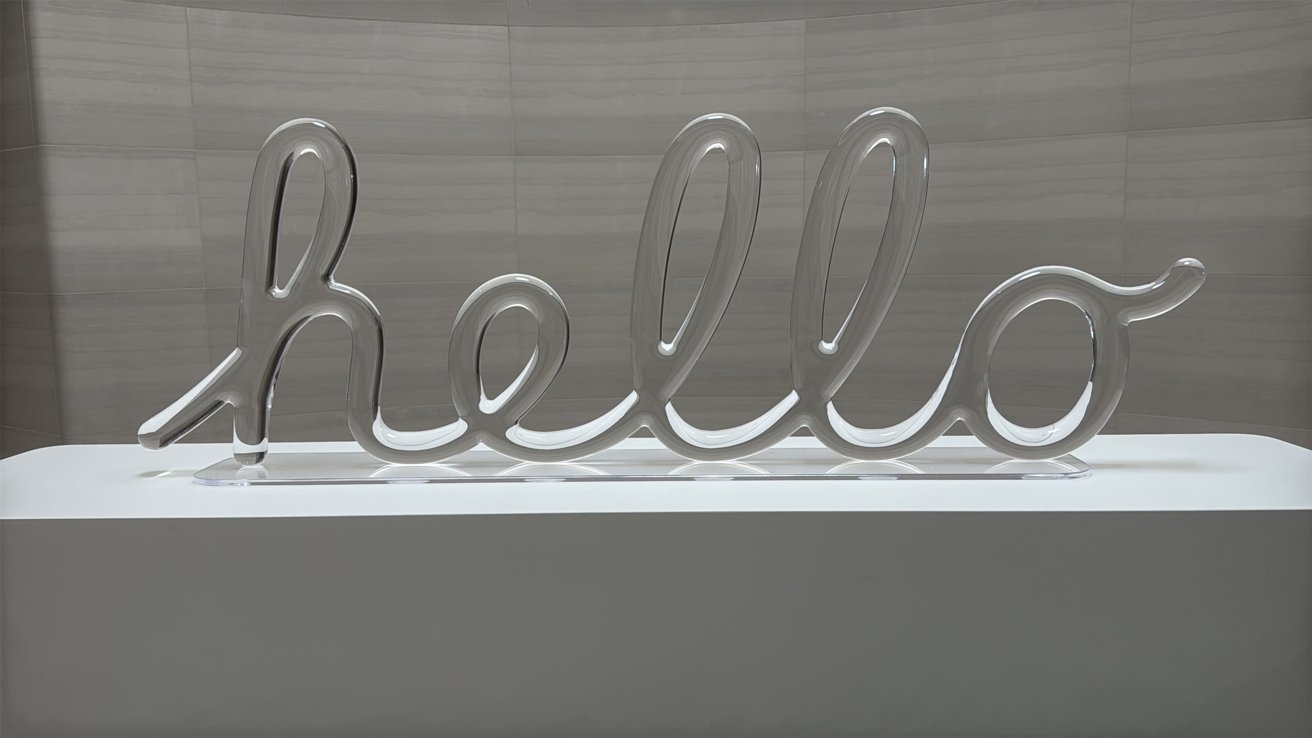 Large clear glass sculpture spelling hello in flowing cursive letters, displayed on a sleek white pedestal against a smooth light gray tiled wall in a modern interior setting