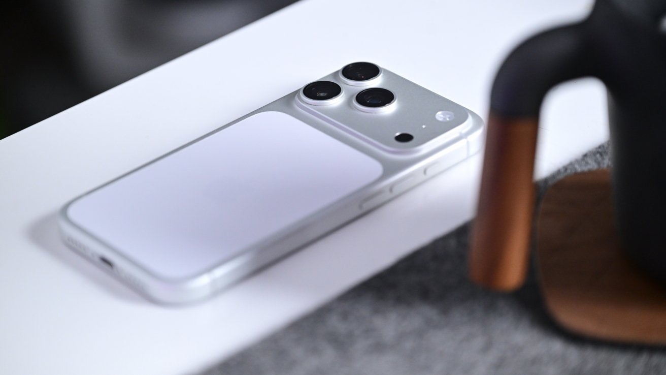 Silver smartphone lying face down on a white surface, showing a large camera module with three lenses and flash, next to a dark mug with a wooden handle on a coaster