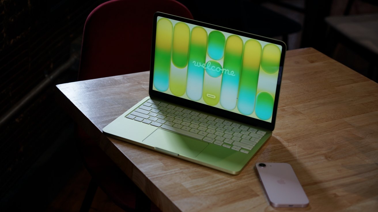 Light green laptop on a wooden table displaying a colorful welcome screen, with a silver smartphone lying nearby; background shows a dark room and a red chair.