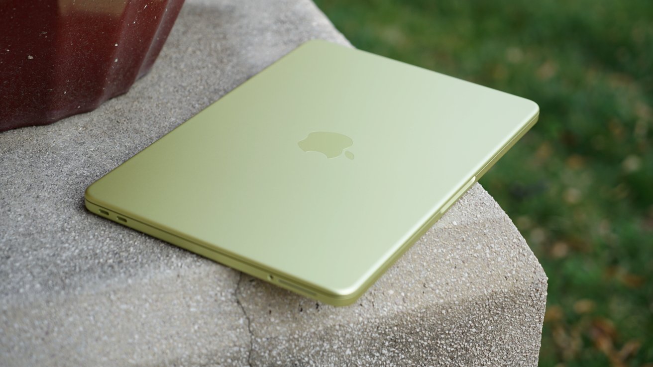 Closed light green Apple MacBook Neo resting on a concrete ledge outdoors, with blurred green grass and scattered leaves in the background