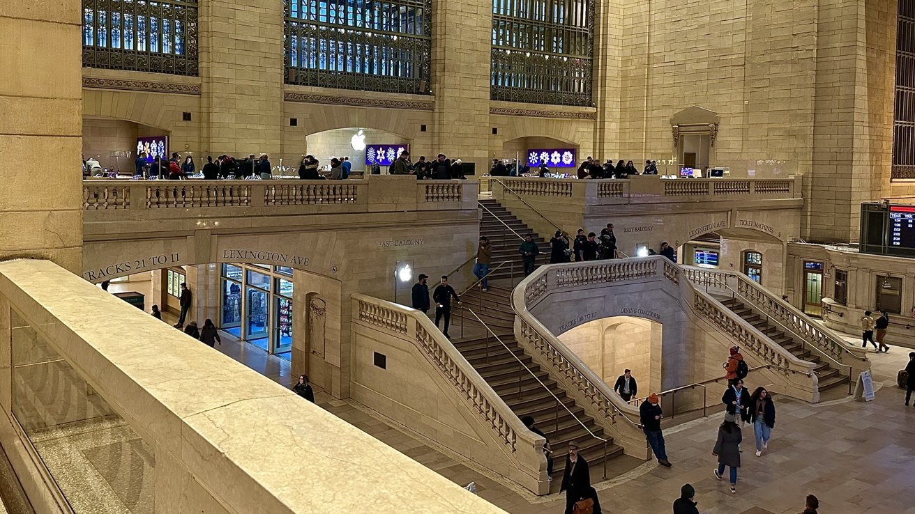 Grand Central Terminal interior with grand staircases, beige stone walls, high windows, people walking and gathering, Apple Store on upper balcony, and entrances leading to tracks and Lexington Avenue