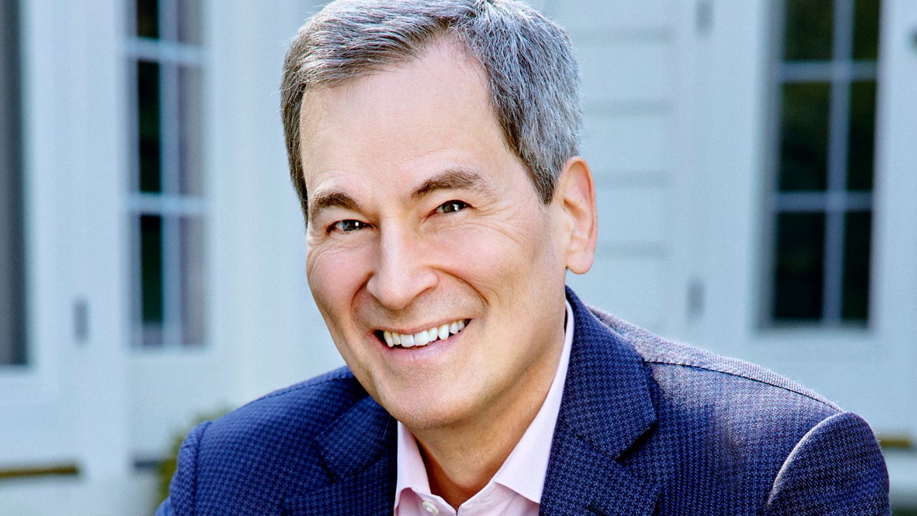 Middleaged man with short gray hair smiling warmly at the camera, wearing a blue blazer and pink shirt, standing outdoors in front of a light-colored house with large windows